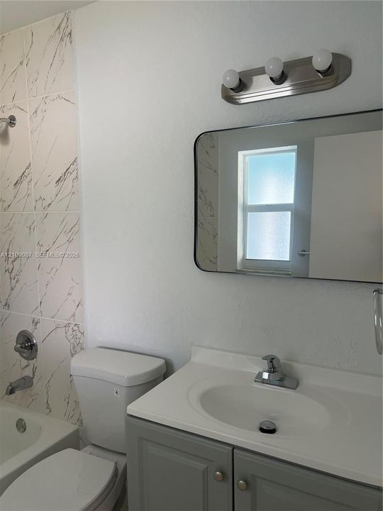 Bathroom featuring a vanity with a white countertop and integrated sink, a rectangular mirror with rounded corners, a three-light vanity fixture, and a bathtub surround with veined tile detailing