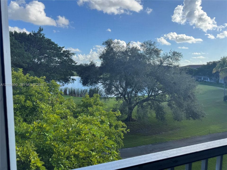 Exterior balcony with a metal railing overlooking a landscape featuring mature trees, a manicured lawn, and a natural pond