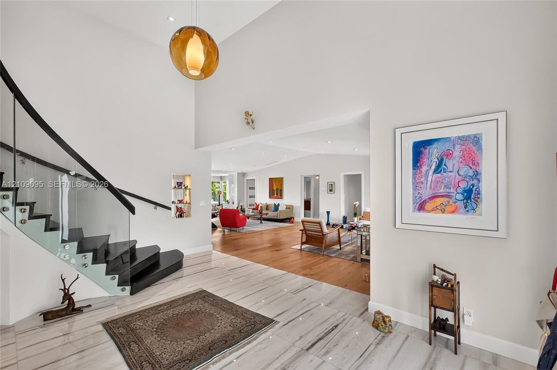 Double-height ceiling, floating staircase with glass paneling and dark-finish treads, stone-style tile flooring, wood-finish flooring, and statement pendant lighting