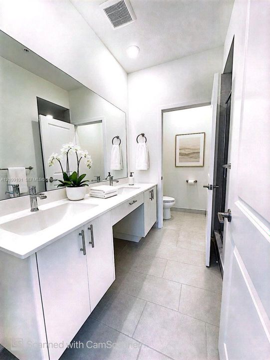 Dual-sink vanity with white cabinetry, sleek hardware, expansive wall-to-wall mirror, and neutral tile flooring