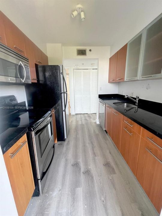 Kitchen featuring wood-finish cabinetry, black stone-look countertops, stainless steel appliances, wood-finish flooring, and frosted glass cabinet accents