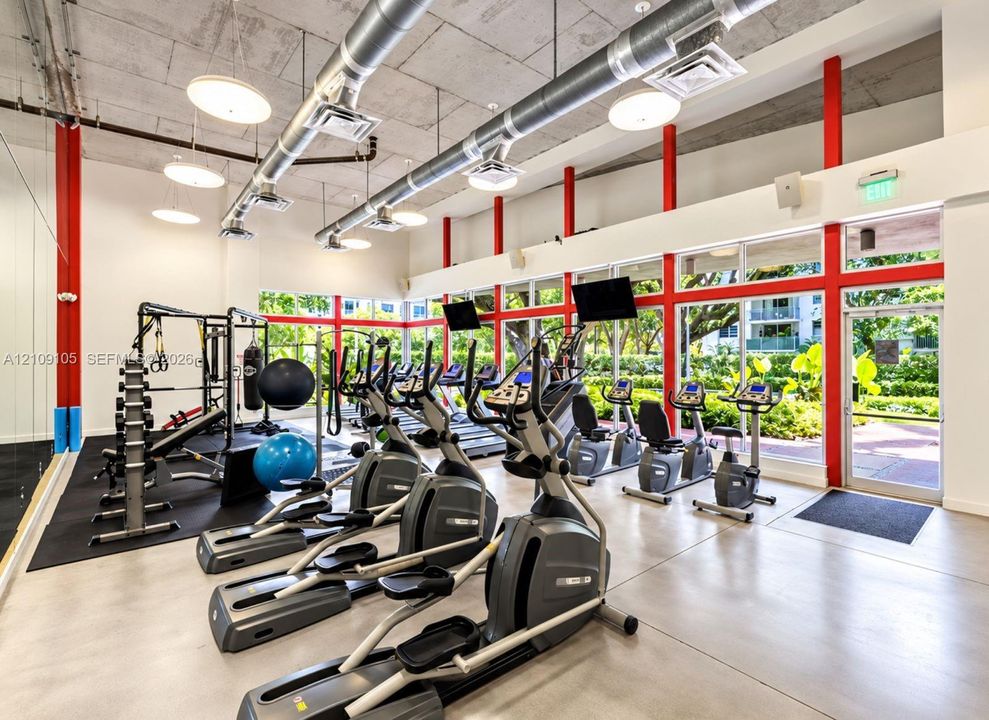 Industrial-style fitness space featuring exposed concrete ceilings, polished concrete flooring, floor-to-ceiling windows with red metal framing, and suspended track lighting