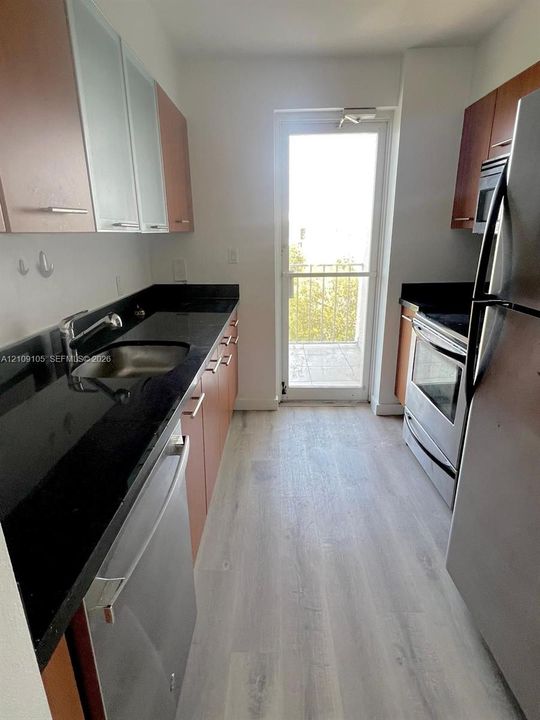 Galley kitchen featuring wood-finish cabinetry, dark solid-surface countertops, stainless steel appliances, and wood-finish flooring with exterior access via a glass-paneled door