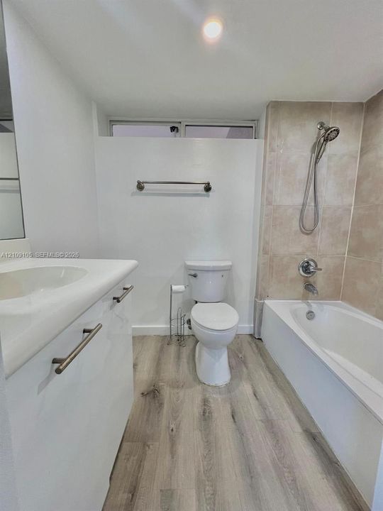 Bathroom featuring a white vanity with an integrated basin, wood-finish flooring, a tub-shower combination with light-toned wall tile, and a rectangular transom window