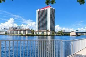 Waterfront high-rise building with a white facade and red roofline, featuring balcony-lined exterior walls and a metal railing along the paved bulkhead