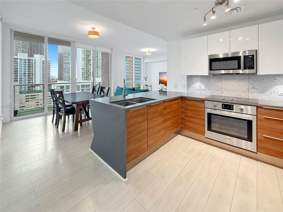 Kitchen featuring a dark solid-surface peninsula, white upper cabinetry, wood-finish base cabinets, stone-patterned backsplash, and light-toned tile flooring