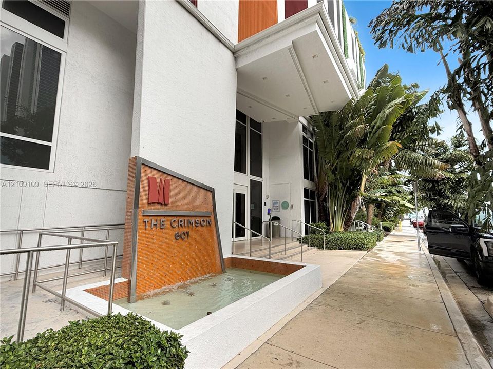 Building exterior featuring a white concrete facade, a prominent illuminated water feature with an integrated metal logo sign, a cantilevered entrance overhang, and lush tropical landscaping