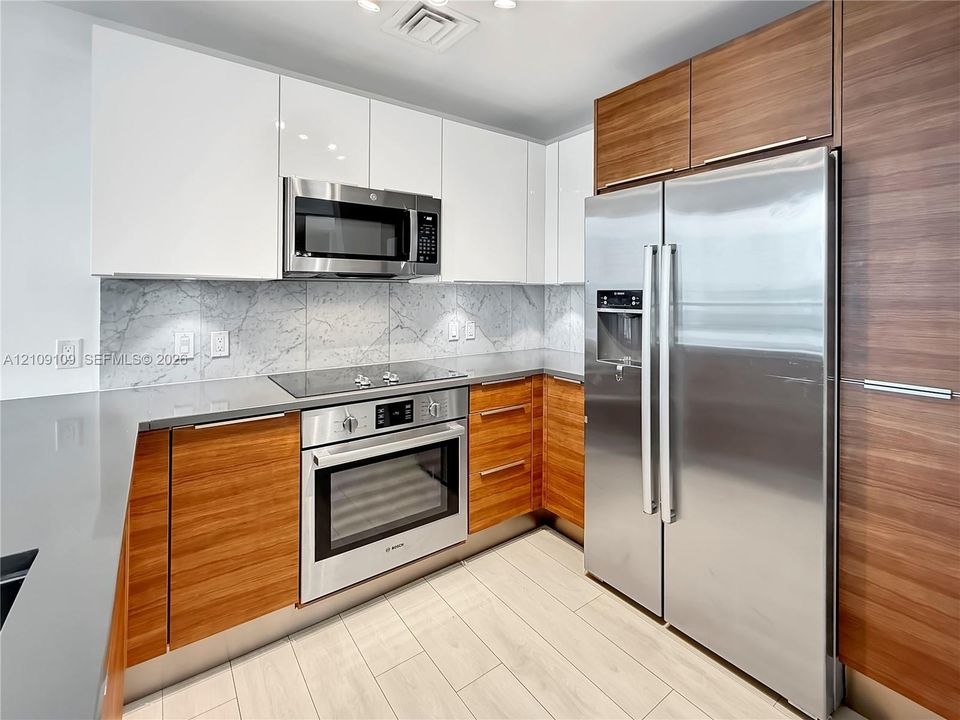 Modern kitchen featuring two-tone cabinetry with horizontal wood grain and matte white finishes, stone-style backsplash, solid-surface countertops, integrated stainless steel appliances, and large-format tile flooring