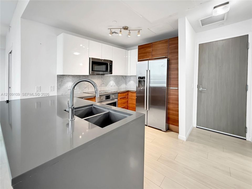 Kitchen featuring a charcoal-toned solid surface island with an undermount double-basin sink, high-gloss white and wood-finish cabinetry, stone-patterned backsplash, and stainless steel appliances