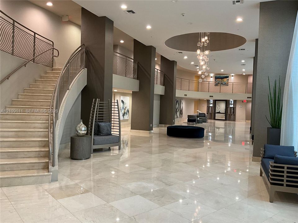 Grand lobby featuring polished stone flooring, a sweeping staircase with custom metal railing, vaulted ceiling with recessed lighting, and a multi-tiered decorative chandelier