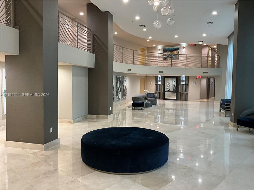 Grand lobby featuring polished stone flooring, a multi-story mezzanine with decorative metal railings, recessed lighting, and a modern glass chandelier