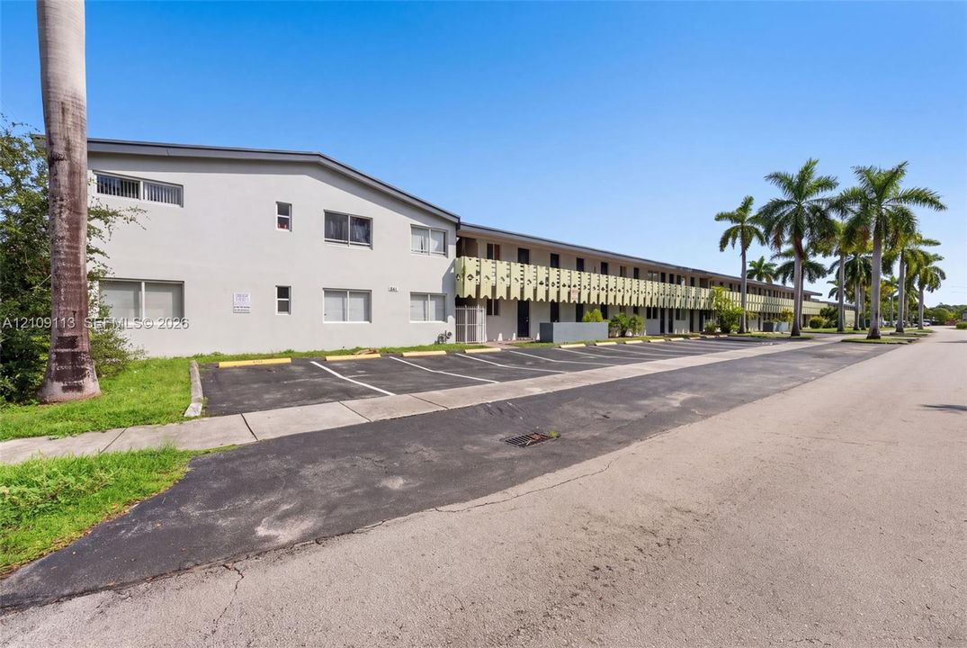 Multi-story exterior featuring a light-toned smooth facade, horizontal window configuration, and dedicated asphalt parking area lined with palm trees