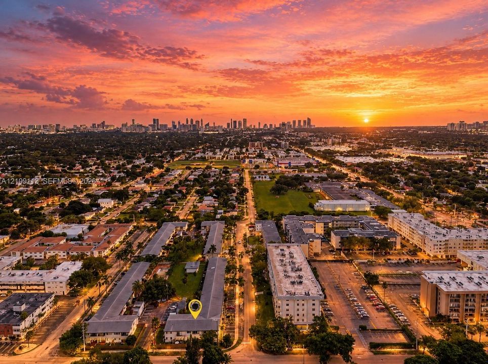 Aerial perspective of a multi-unit residential complex featuring flat roofs, exterior walkways, and mature landscaping, complemented by a sunset horizon and city skyline backdrop