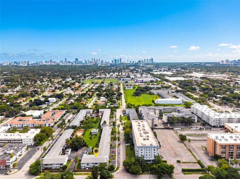Aerial perspective featuring a multi-unit residential complex with surface parking, manicured landscaping, palm trees, and prominent city skyline backdrop