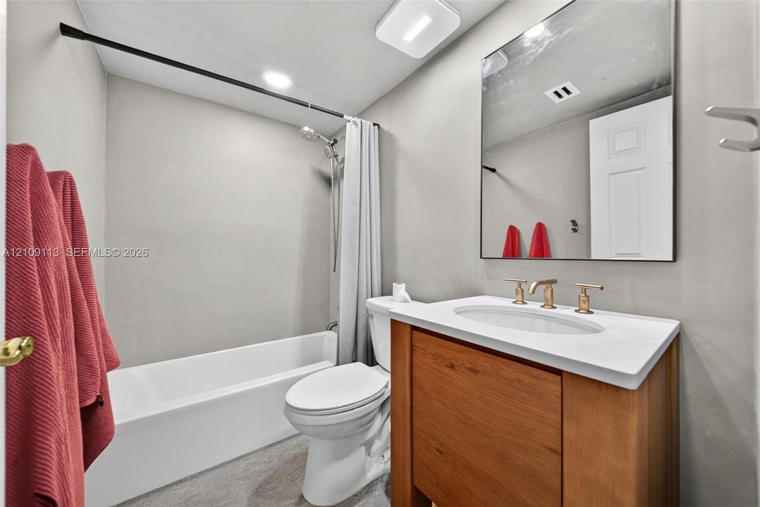 Bathroom featuring a wood-finish vanity with a stone-top integrated sink, brushed gold-tone faucet, rectangular frameless mirror, and a deep soaking tub with a wall-mounted shower fixture