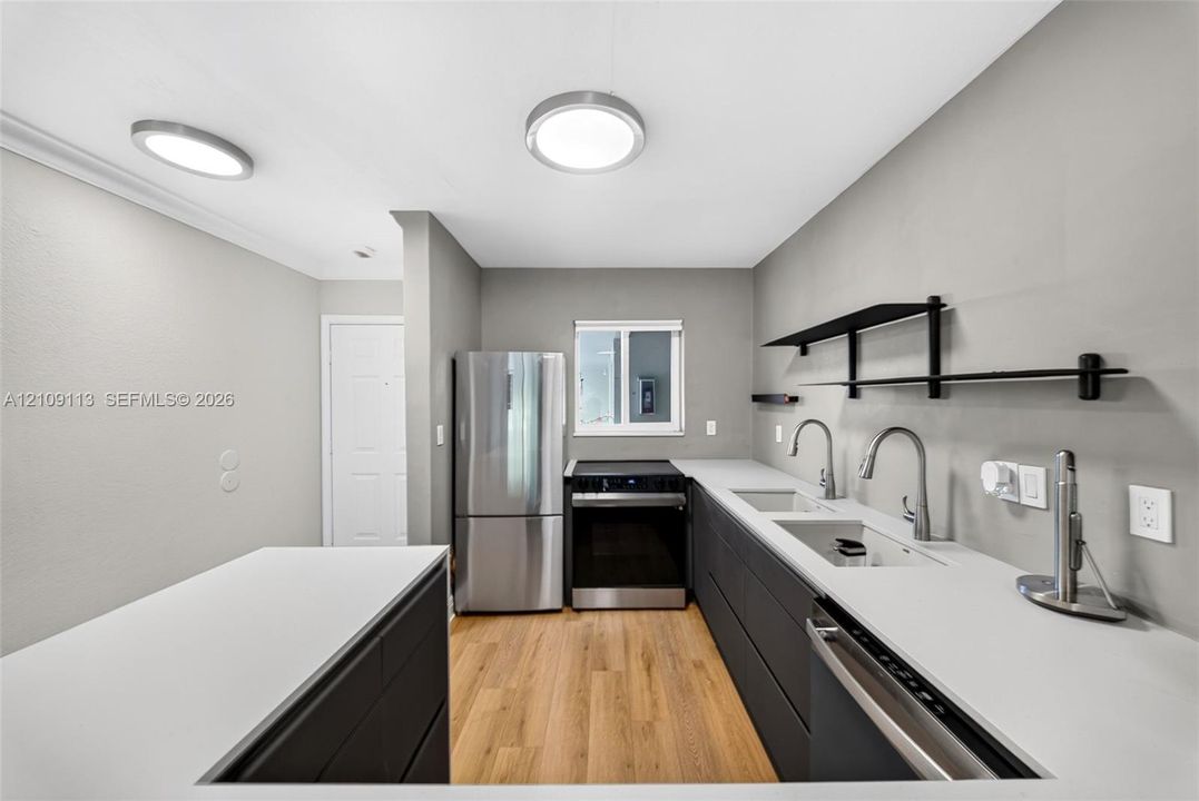 Modern kitchen featuring dark cabinetry, solid-surface countertops, integrated stainless steel appliances, wood-finish flooring, and black wall-mounted shelving