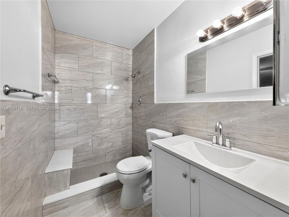 Bathroom featuring large-format tile wall surrounds, a walk-in shower with a built-in bench, a white vanity with integrated basin, and polished chrome fixtures