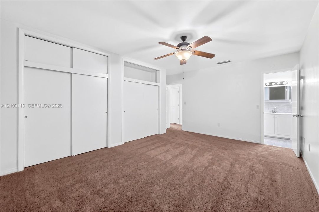 Room with neutral wall-to-wall carpeting, dual sliding-door closets with transom windows, and a ceiling fan with integrated lighting
