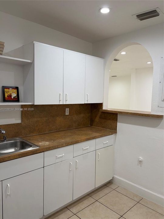 White cabinetry, laminate countertops, stainless steel sink, arched opening with a pass-through ledge, and tile flooring