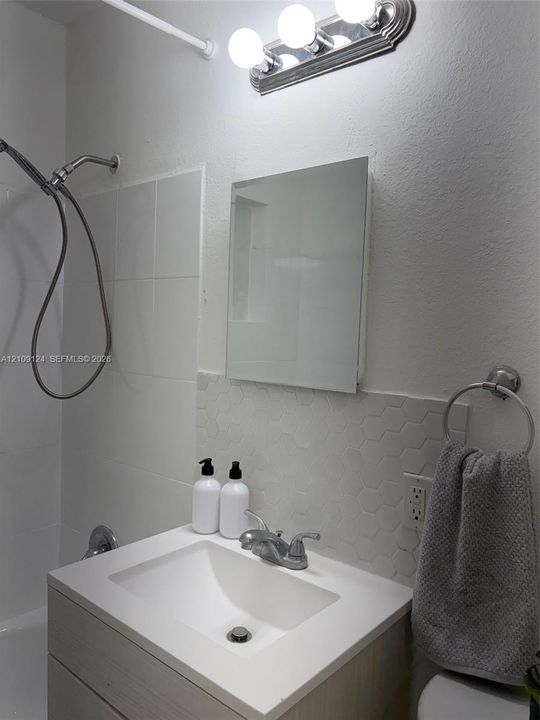 Vanity with a white rectangular basin, light wood-finish cabinetry, hexagonal tile backsplash, wall-mounted mirror, and a multi-bulb vanity light fixture