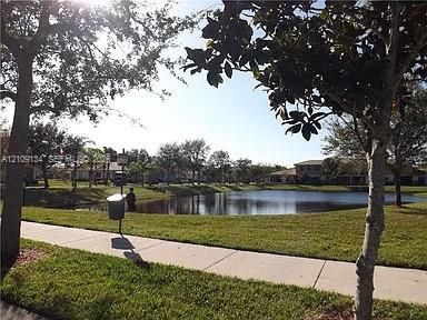 Waterfront grounds featuring a concrete sidewalk, expansive lawn, mature trees, and a pond