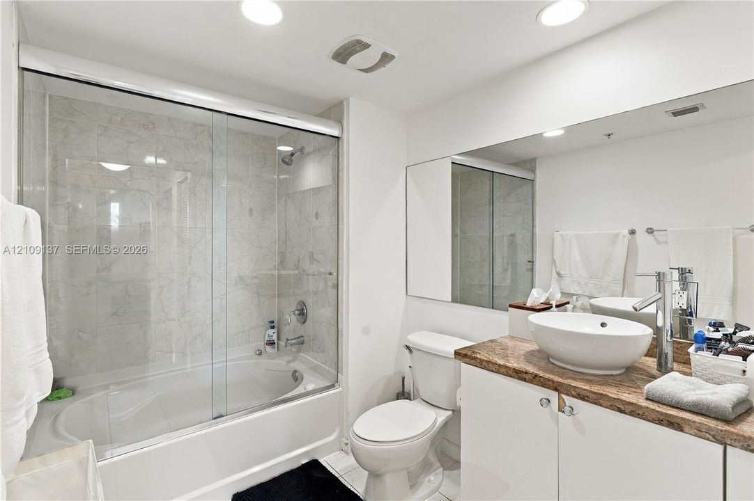 Bathroom featuring a vessel sink atop a stone-finish countertop, white cabinetry, a full-width mirror, and a soaking tub with a sliding glass enclosure and tiled surround
