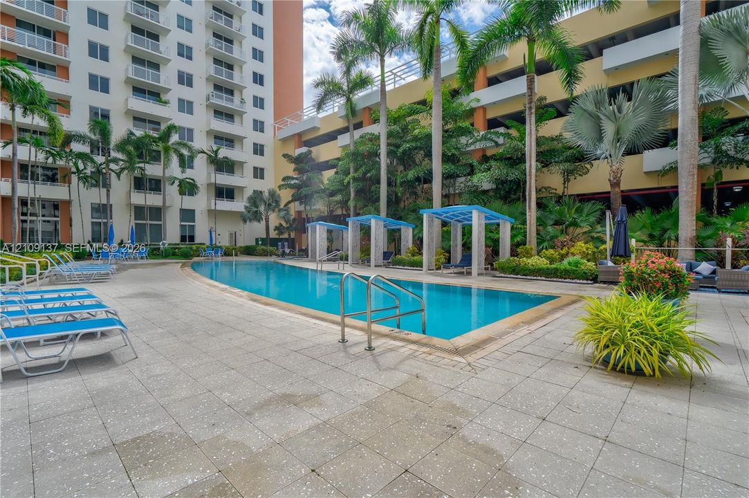 Curved swimming pool with metal handrails, paved pool deck, and shaded pergola structures set among palm trees