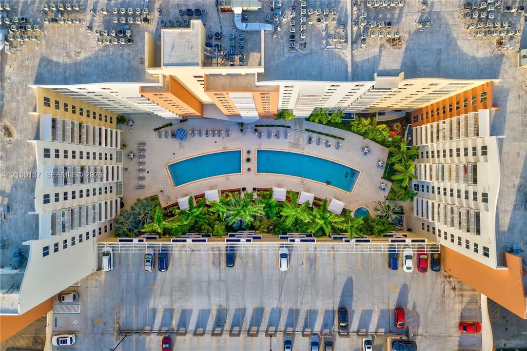 Aerial perspective of a multi-story residential complex featuring a central pool area, surrounding palm landscaping, hardscaped patio space, and dedicated surface parking