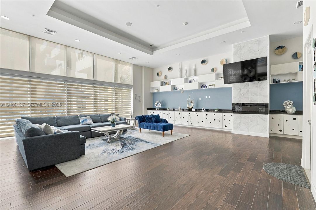 Expansive living space featuring wood-finish flooring, floor-to-ceiling windows with roller shades, recessed tray ceilings, built-in cabinetry, and a fireplace with stone-finish surround