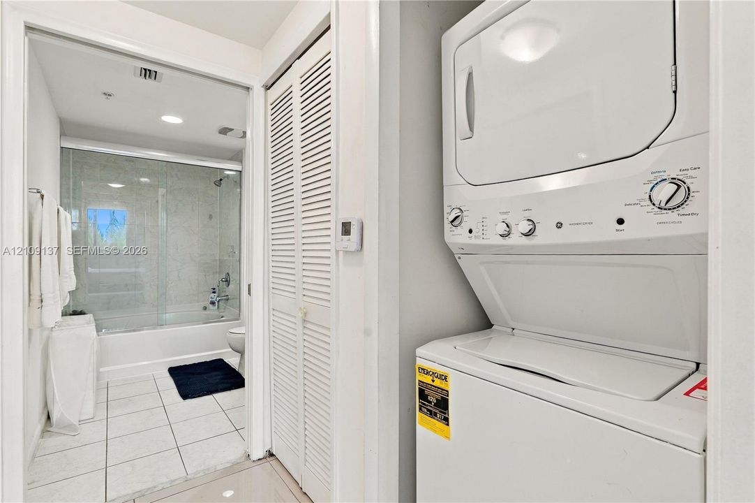 Stacked laundry unit with white finish, closet with louvered doors, bathroom featuring a tub-shower combination with a glass enclosure, and ceramic tile flooring