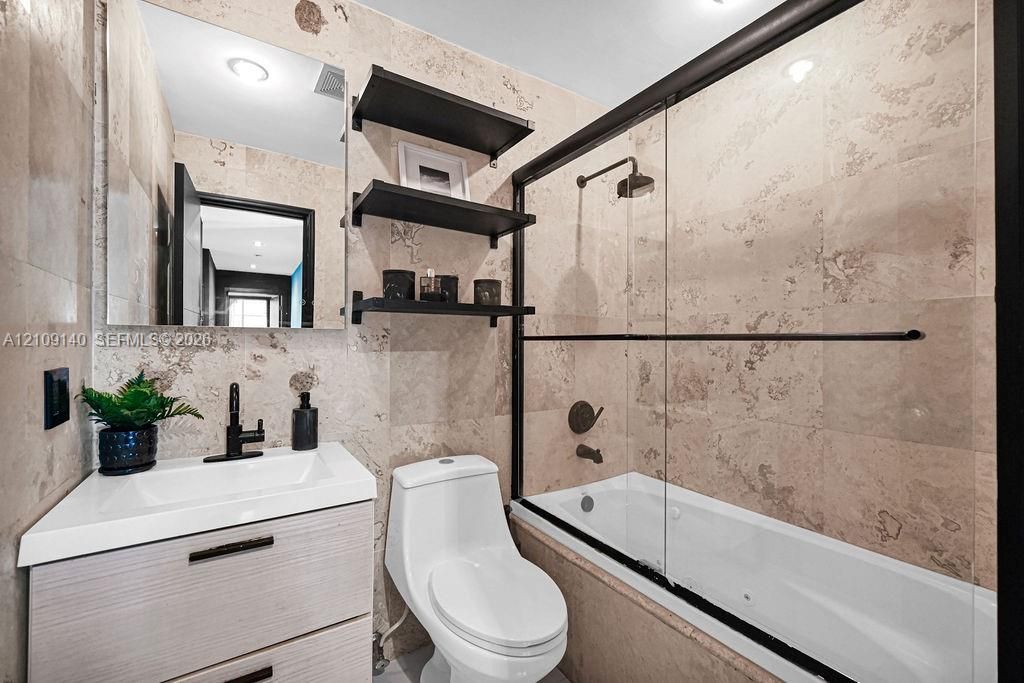 Bathroom featuring floor-to-ceiling stone tile, a modern vanity with integrated sink, matte black hardware, floating shelving, and a glass-enclosed soaking tub with a shower head