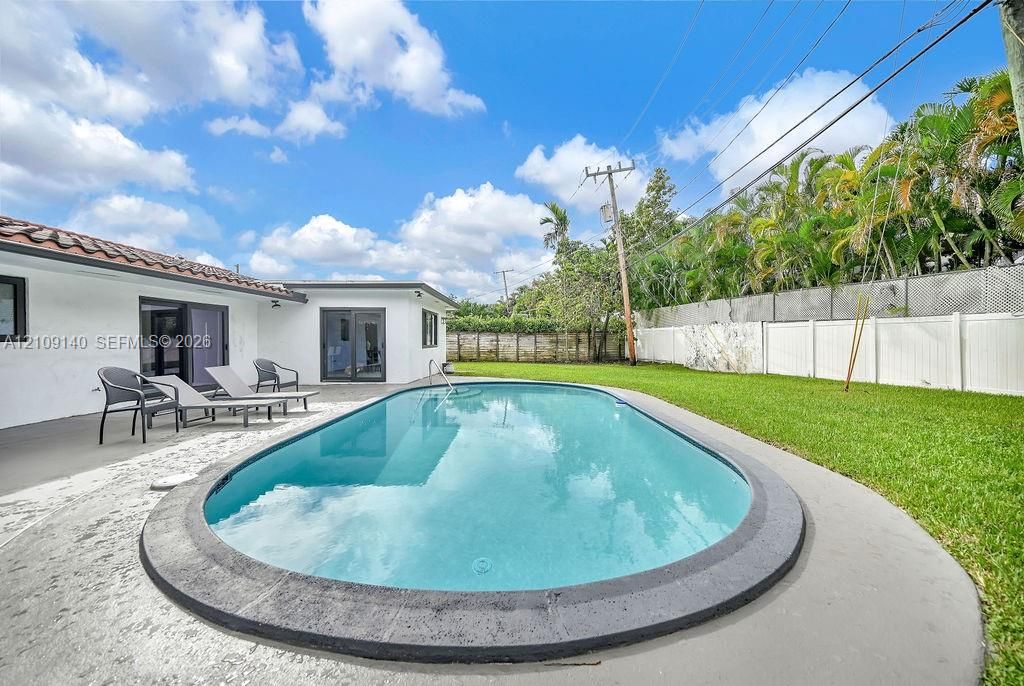 Oval-shaped swimming pool with a dark stone-finish coping, concrete patio, white exterior wall finish, terra cotta roof accents, and a grass lawn bounded by privacy fencing