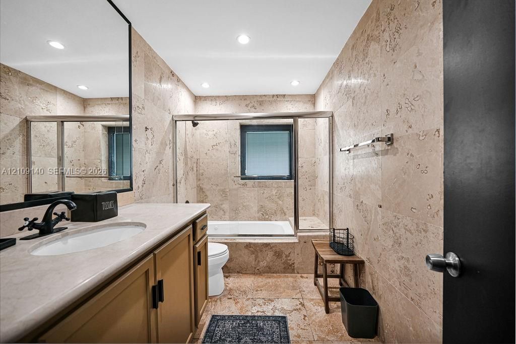 Full-height stone wall tiling, integrated bathtub and shower enclosure, stone countertop vanity with matte black faucet, and recessed lighting