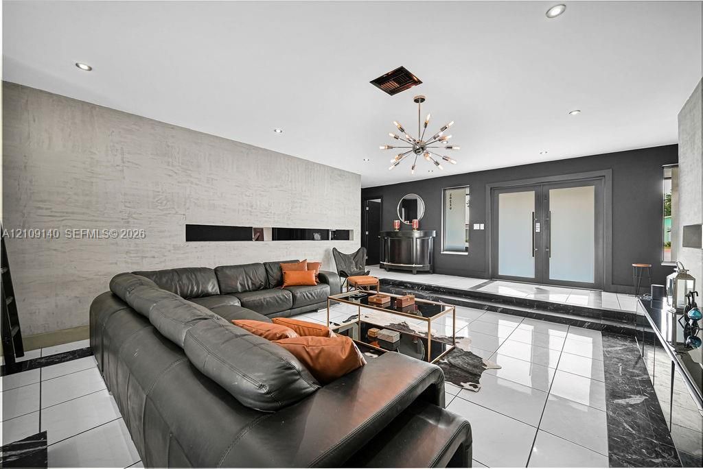 Spacious interior featuring large-format tile flooring with dark stone inlays, a textured concrete feature wall, dual-entry doors with frosted glass, recessed lighting, and a modern multi-arm sputnik chandelier