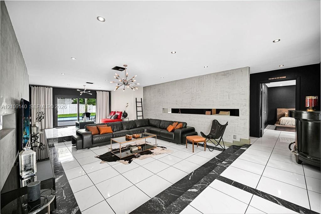 Expansive living space featuring large-format white tile flooring with dark stone inlay borders, a textured concrete accent wall, recessed lighting, and a modern sputnik-style chandelier