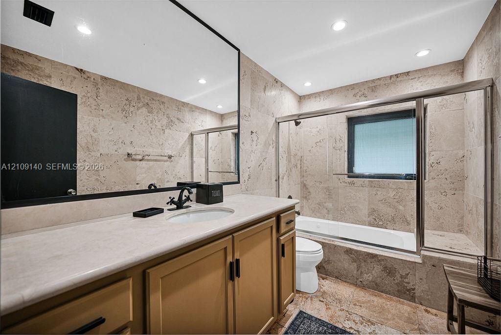 Bathroom featuring full-wall stone tiling, built-in vanity with a solid surface countertop, undermount sink, matte black hardware, and a glass-enclosed bathtub with a shower head