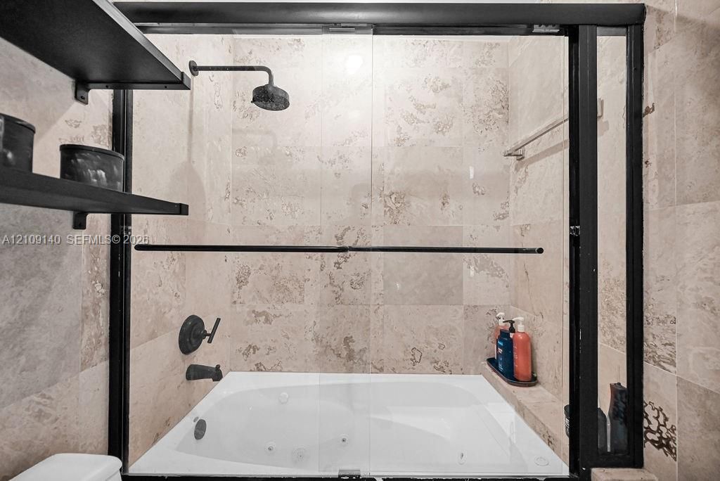 Jetted soaking tub with integrated shower head, black metal framed glass enclosure, stone-tile wall surround, and mounted black shelving