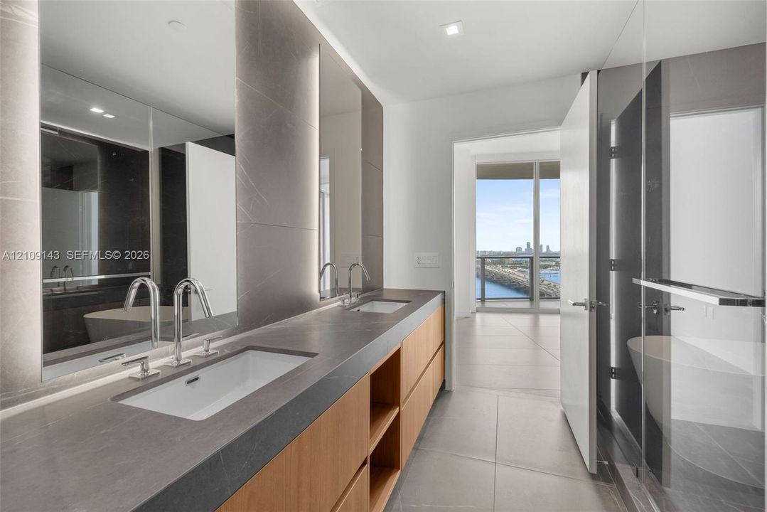 Dual-sink vanity with a solid-surface countertop, wood-finish cabinetry, floor-to-ceiling tile mirror surrounds, glass-enclosed shower, and large-format tile flooring