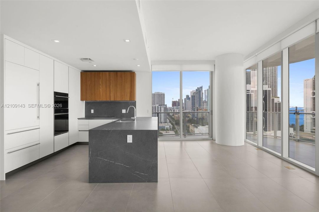 Modern kitchen featuring matte white cabinetry, wood-finish upper cabinets, a dark stone-finish waterfall island, recessed lighting, and floor-to-ceiling windows