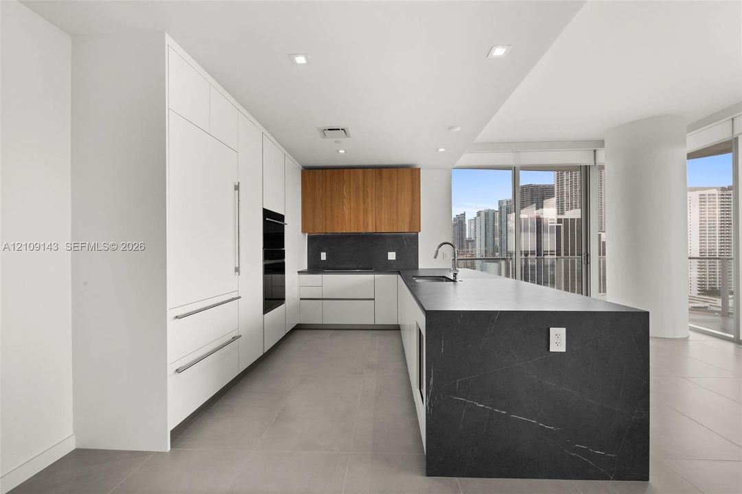 Kitchen featuring flat-panel white cabinetry, wood-finish upper cabinets, expansive waterfall-edge stone island, built-in wall oven, and large-format tile flooring