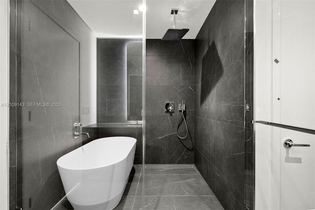 Freestanding soaking tub, large-format dark tile wall surfacing, overhead rainfall showerhead, handheld shower fixture, and frameless glass enclosure