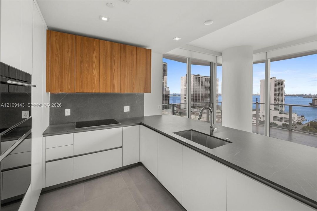 Kitchen featuring wood-finish upper cabinetry, matte white base cabinetry, dark stone-finish countertops and backsplash, stainless steel undermount sink, and floor-to-ceiling windows