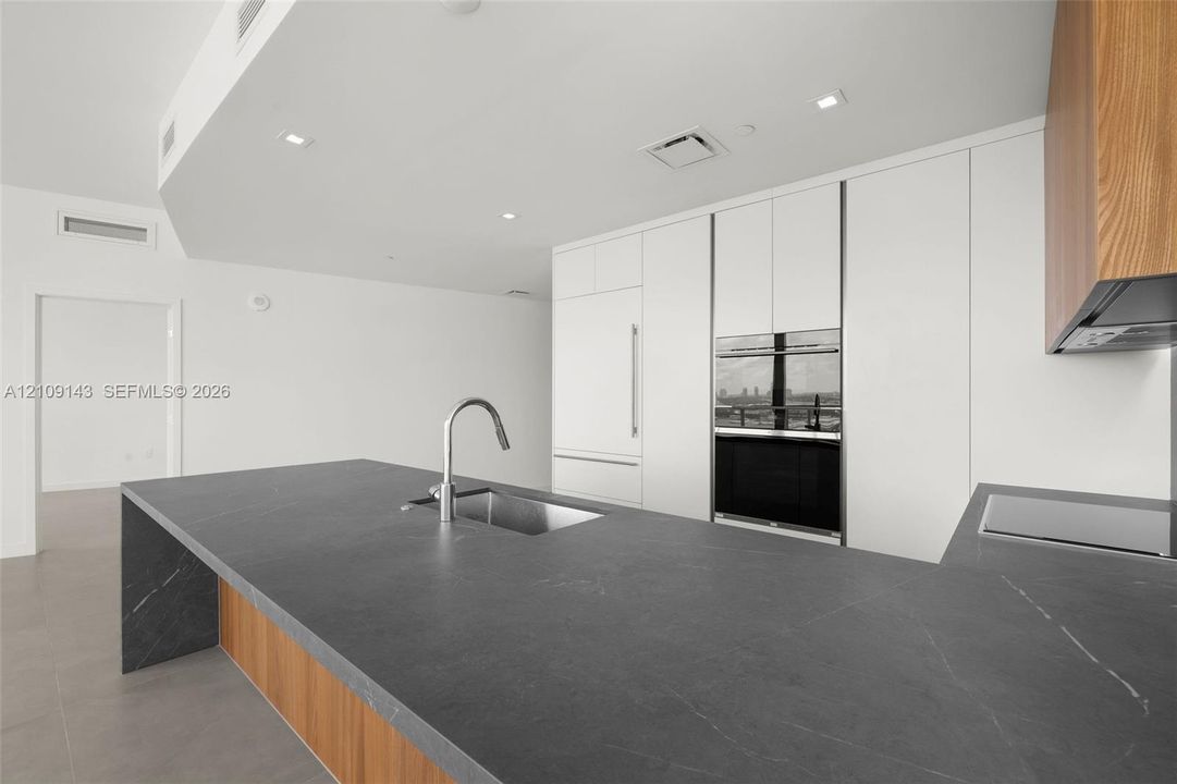 Kitchen featuring a waterfall-edge island with dark stone countertop, undermount stainless steel sink, flat-panel white cabinetry, wood-finish lower cabinetry, and integrated stainless steel wall ovens
