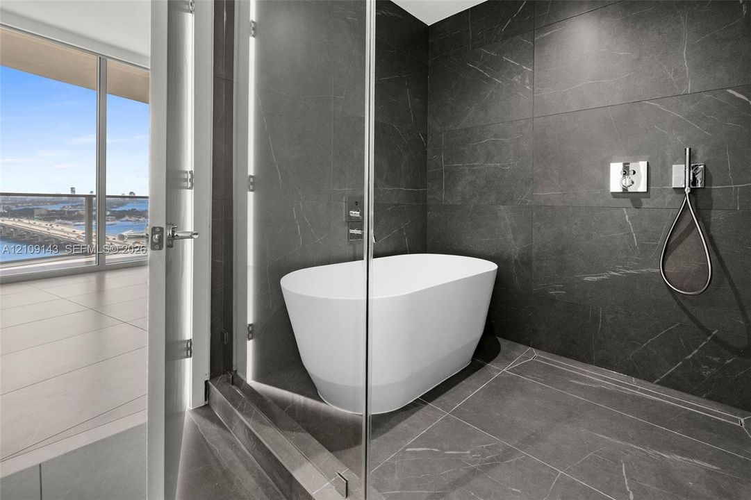 Freestanding soaking tub set within a glass-enclosed wet room featuring floor-to-ceiling dark-toned stone-finish tile and polished chrome plumbing fixtures