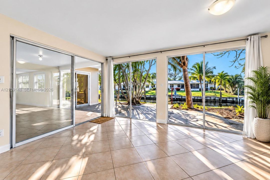 Expansive room featuring wall-to-wall sliding glass doors, neutral tile flooring, and direct access to a waterfront patio with palm trees