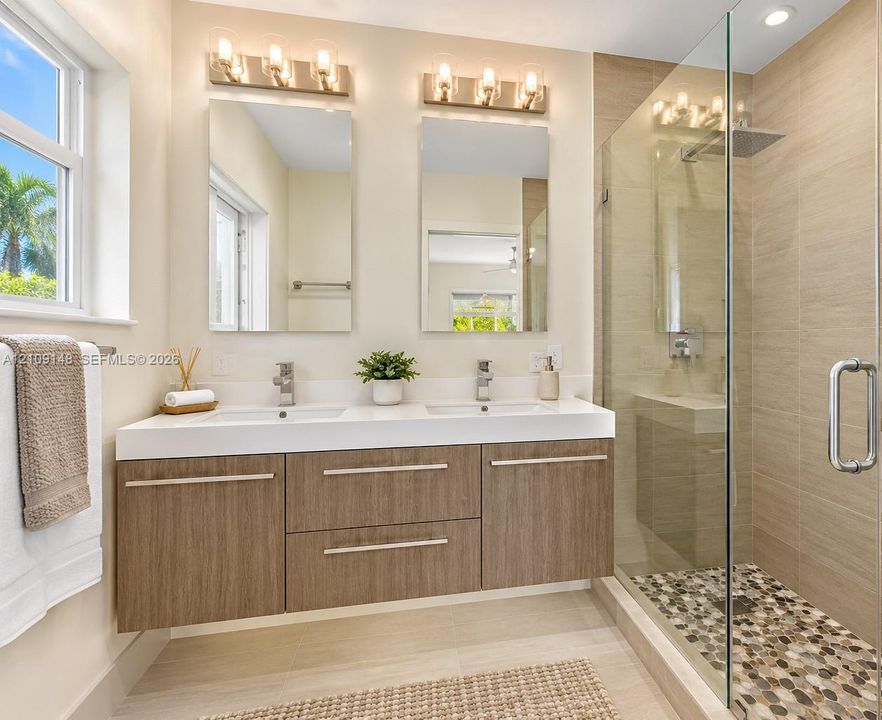Dual-sink vanity with wood-finish cabinetry, quartz countertop, framed glass shower enclosure, stone pebble shower flooring, and wall-mounted vanity lighting