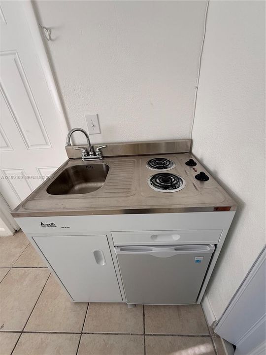 Compact kitchenette unit featuring a stainless steel integrated sink and dual-burner electric cooktop, white cabinetry, and a built-in under-counter refrigerator