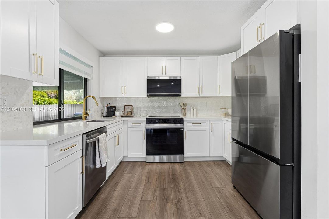 Kitchen featuring shaker-style white cabinetry, quartz countertops, stainless steel appliances, brass-finish hardware, and wood-finish flooring