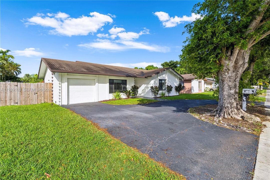 Single-story exterior featuring white stucco siding, a low-pitch roof, an attached one-car garage, asphalt driveway, and a wood-panel fence