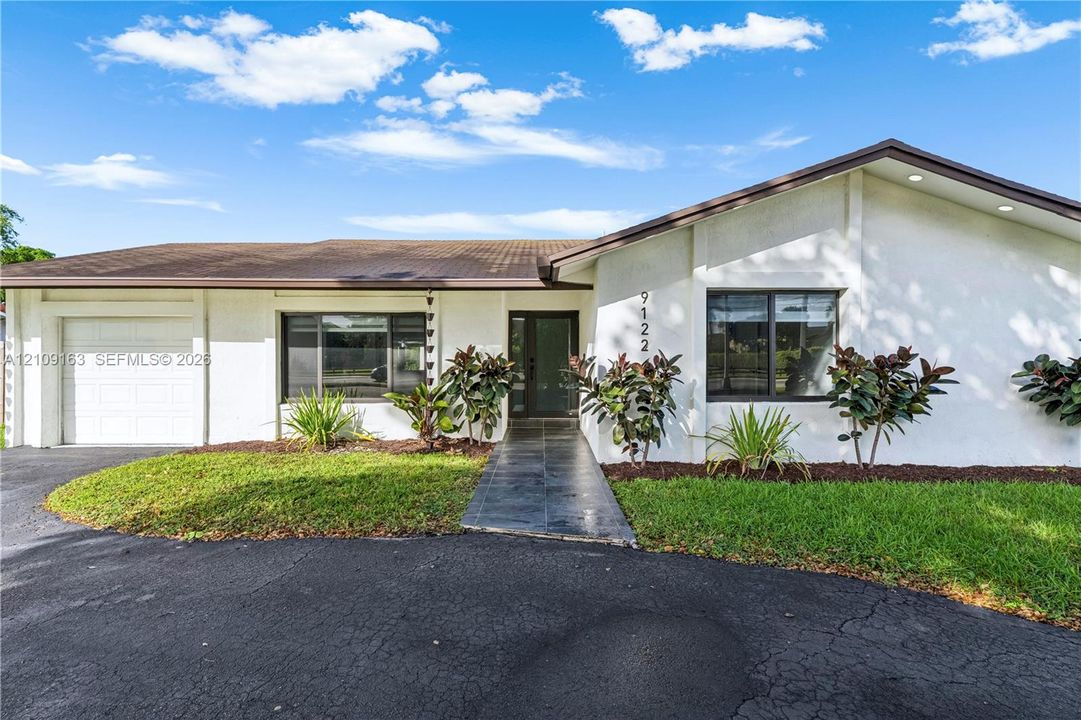 Single-story exterior featuring white stucco siding, a tiled roof, a paved walkway, a multi-pane glass entry door, and an attached garage